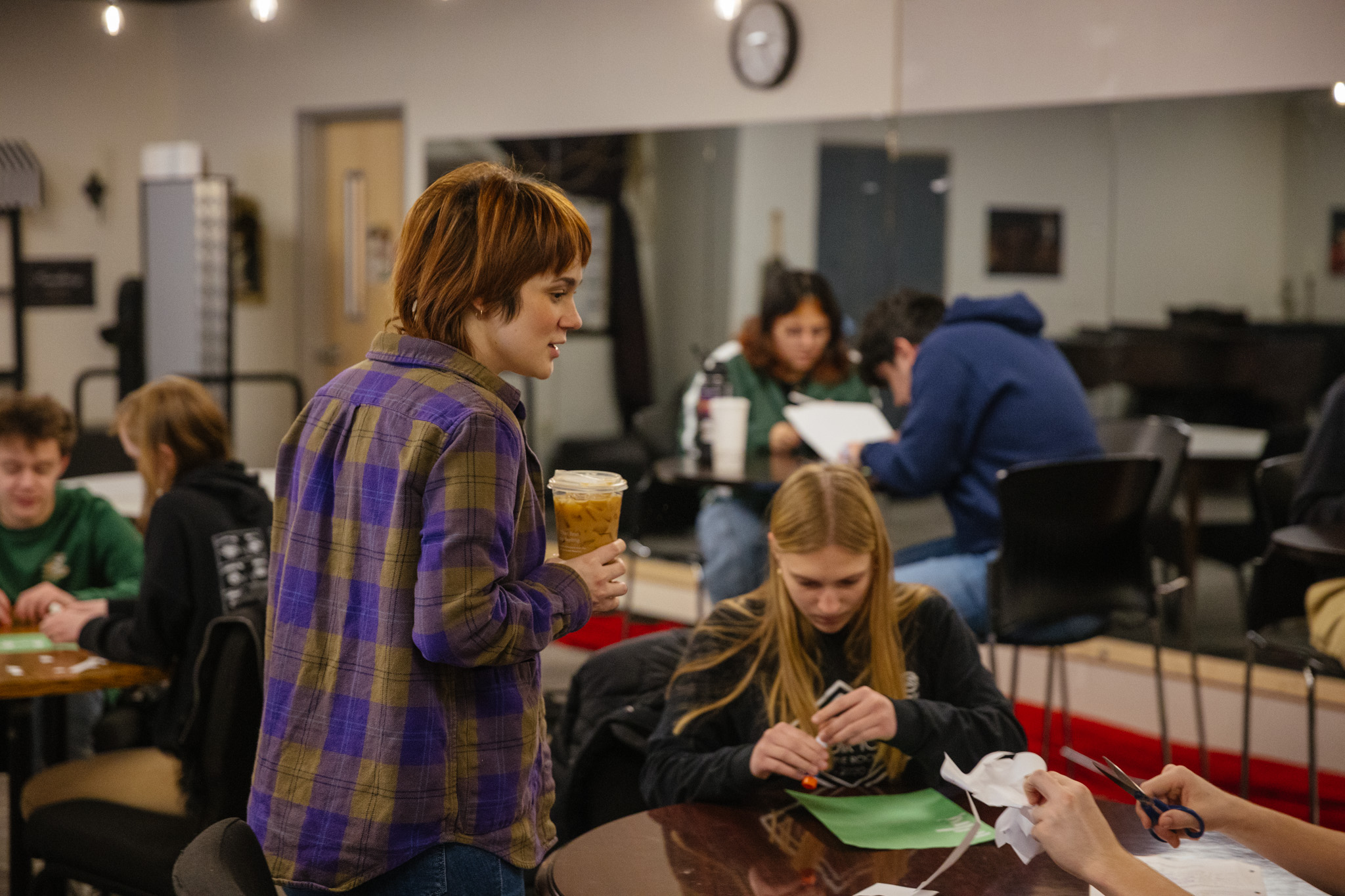 A high school student wearing a purple plaid shirt instructs students while standing in a classroom at New Life Academy. Other students are seated around tables, engaged in various activities such as cutting paper and studying, creating a lively and collaborative learning environment.