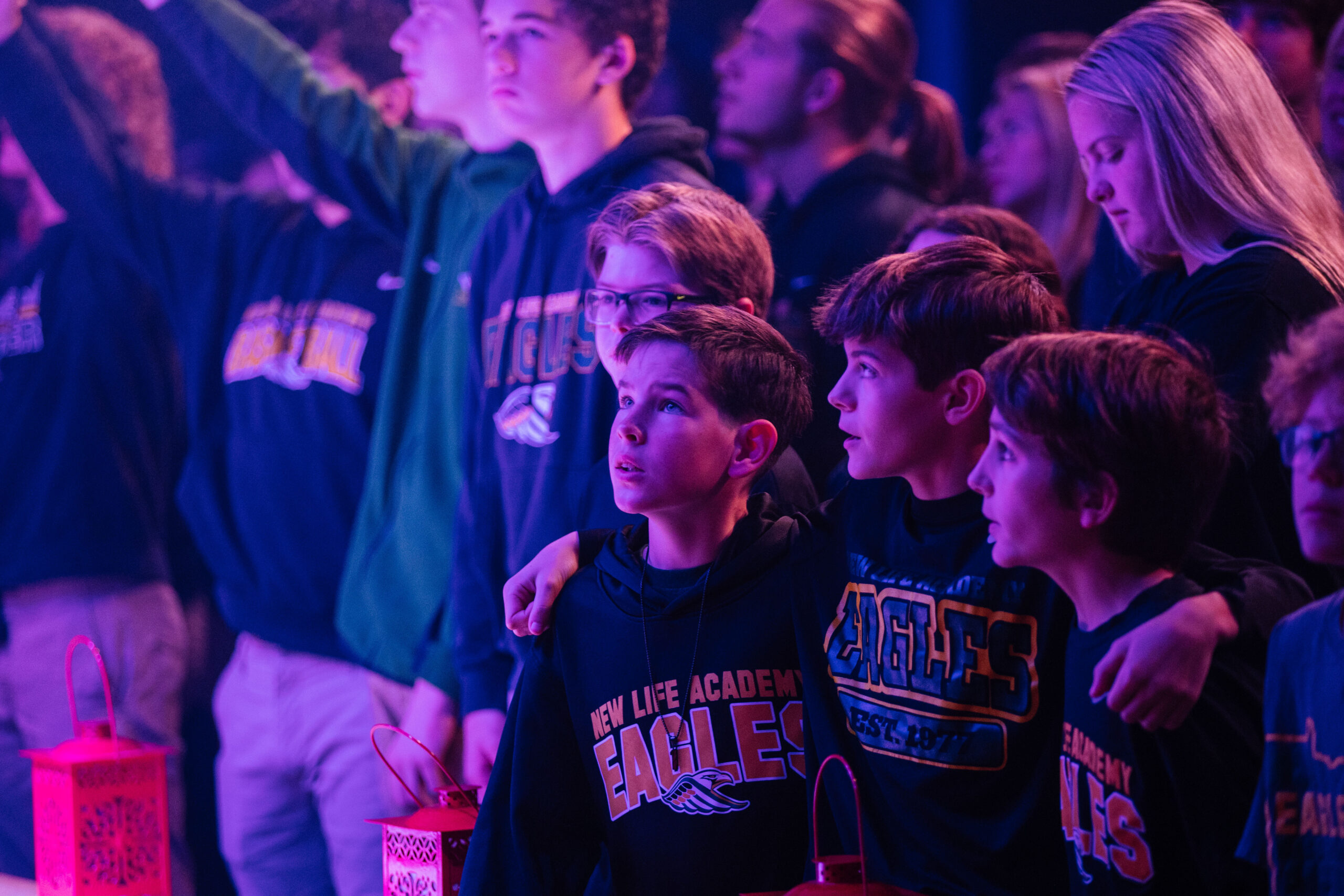 Three Middle School students in New Life Academy sweatshirts smile at the camera outside with fall grass and trees behind them.